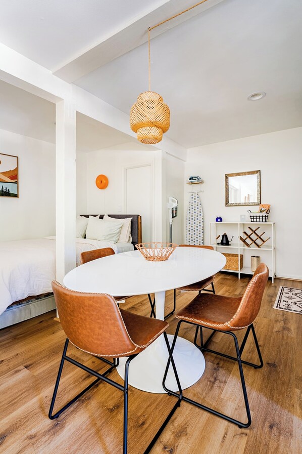A modern room with a round dining table, two brown leather chairs, a woven pendant light, a bed with white bedding, and a white wall with a framed mirror and shelves.