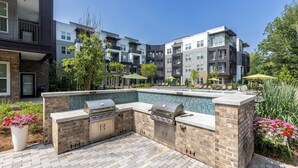 An outdoor pool area with a barbecue setup, surrounded by a brick patio and lush greenery. - Landing | Modern 1BD, Yoga Studio, Pool (Charlotte)