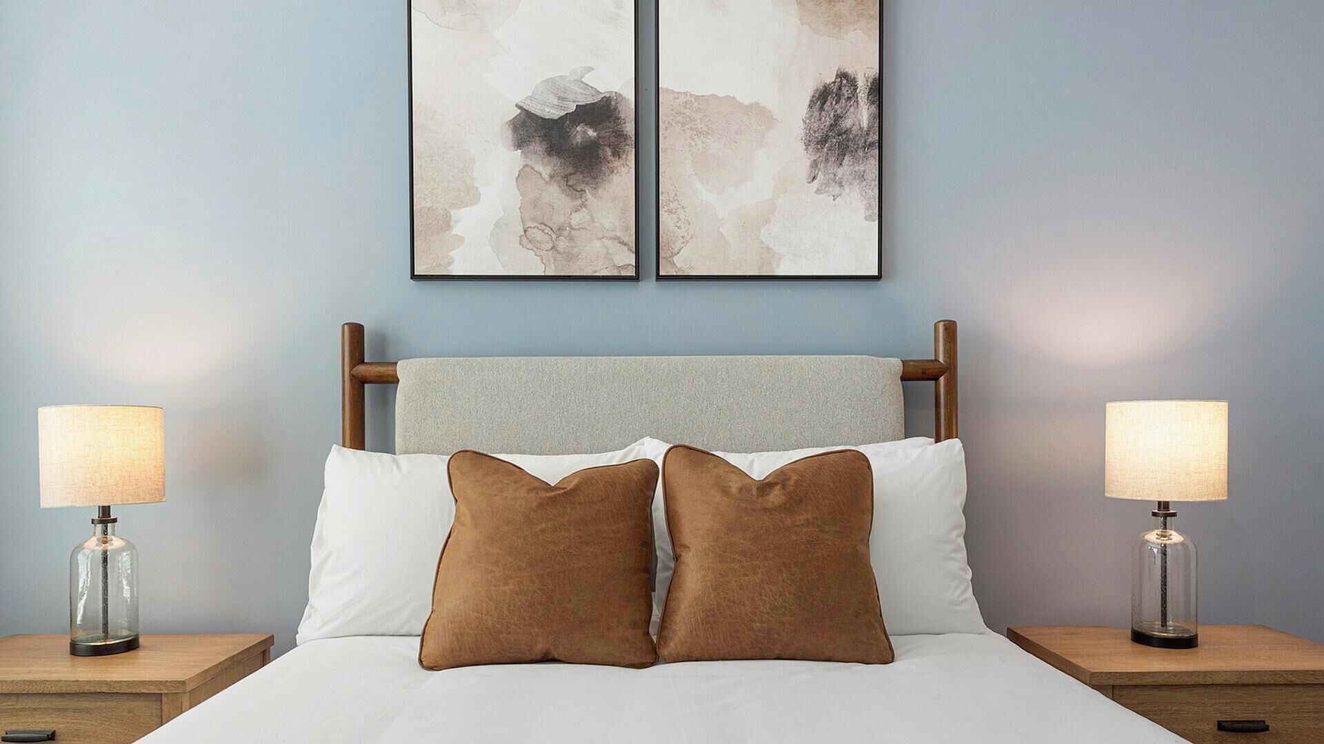 Uma cama com cabeceira de madeira, dois travesseiros castanhos, lençóis brancos e duas luminárias de cabeceira em mesas de apoio de madeira.