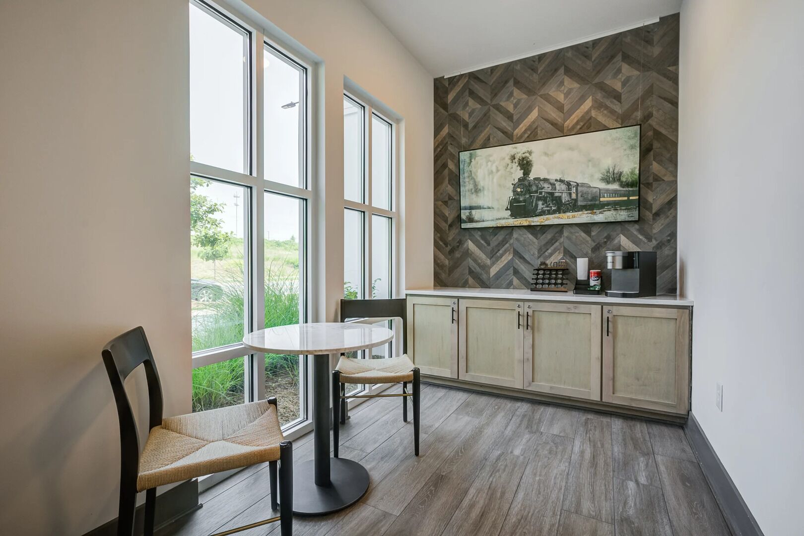 A modern kitchen with large windows, a dining area with a round table and chairs, and a herringbone-patterned backsplash above the cabinets.