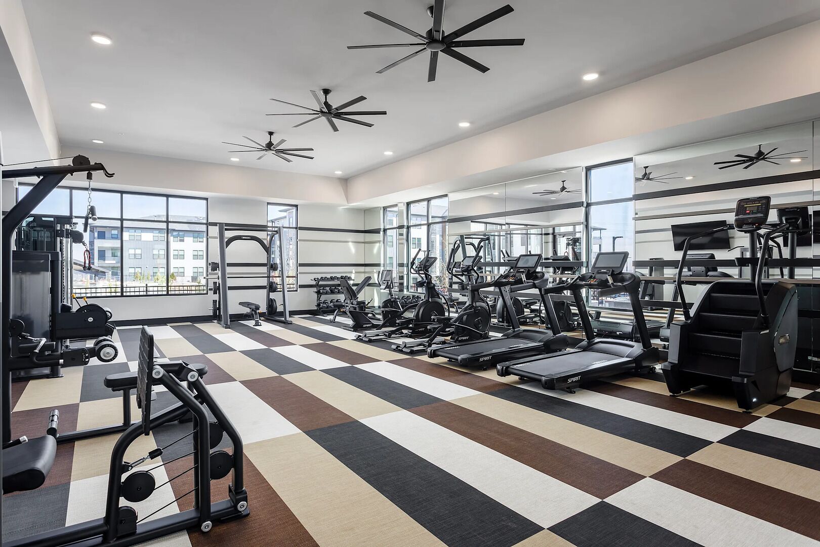 Une salle de sport équipée de divers appareils d’exercice, dotée de grandes fenêtres et de ventilateurs de plafond.