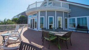 Een overdekt terras buiten met een eettafel en stoelen, een witte balustrade en een gebogen gebouw met grote ramen.