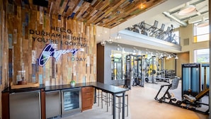 A gym with exercise equipment, a wooden wall with a neon sign, and a mezzanine level with seating. - Landing | Beautiful 1BD, Game Room (Houston)