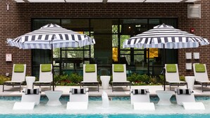 A poolside area with lounge chairs and striped umbrellas, a brick building facade, and a clear view of the pool. - Landing | Beautiful 1BD, Game Room (Houston)