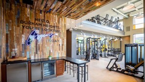 A gym with exercise equipment, a wooden wall with a neon sign, and a mezzanine level with seating. - Landing | Spectacular 1BD, Game Room (Houston)