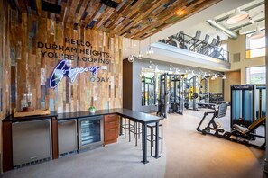 Ein Fitnessraum mit Trainingsgeräten, eine Holzwand mit einem Neonzeichen und eine erhöhte Ebene mit Sitzgelegenheiten.
