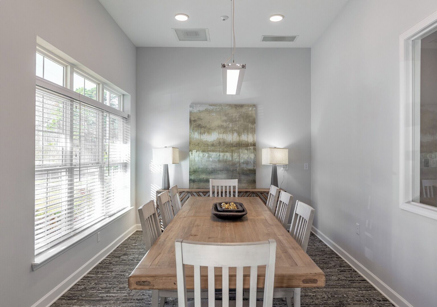 A dining room with a wooden table, white chairs, two floor lamps, and a large abstract painting on the wall.