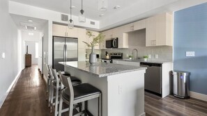 A modern kitchen with a central island, bar stools, stainless steel appliances, and a blue accent wall. - Landing | Spectacular 1BD, Clubhouse (Austin)