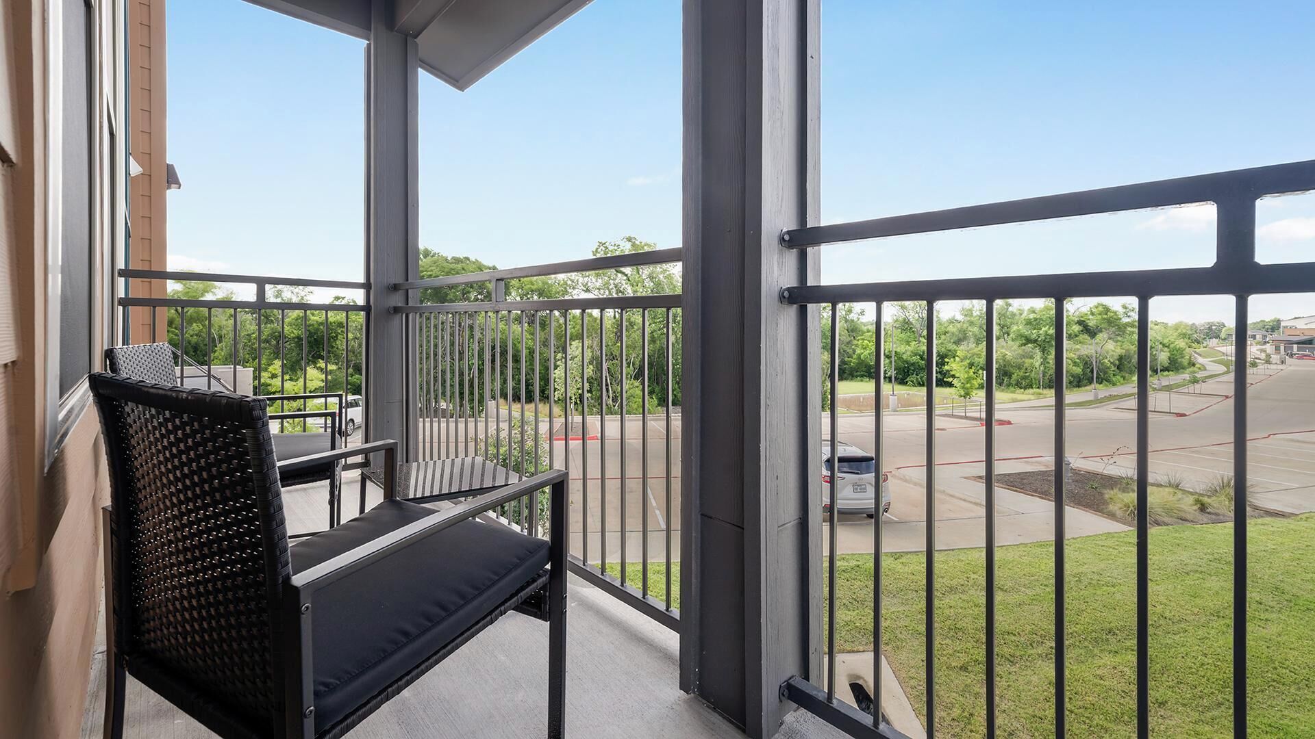A balcony with a black chair and a view of a parking lot and greenery.