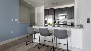 A modern kitchen with dark cabinets, a stainless steel refrigerator, a microwave, and a stove. There are three bar stools and a small potted plant on the counter. - Landing | Incredible 1BD, Gym, Pool (Austin)