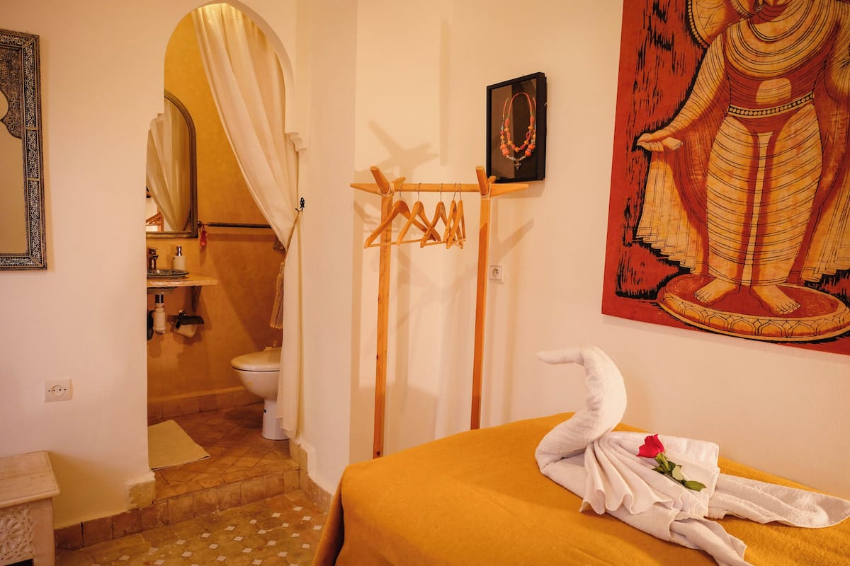 A room with a bed, a towel with a rose, a painting, a wooden rack, and a bathroom visible through an archway.