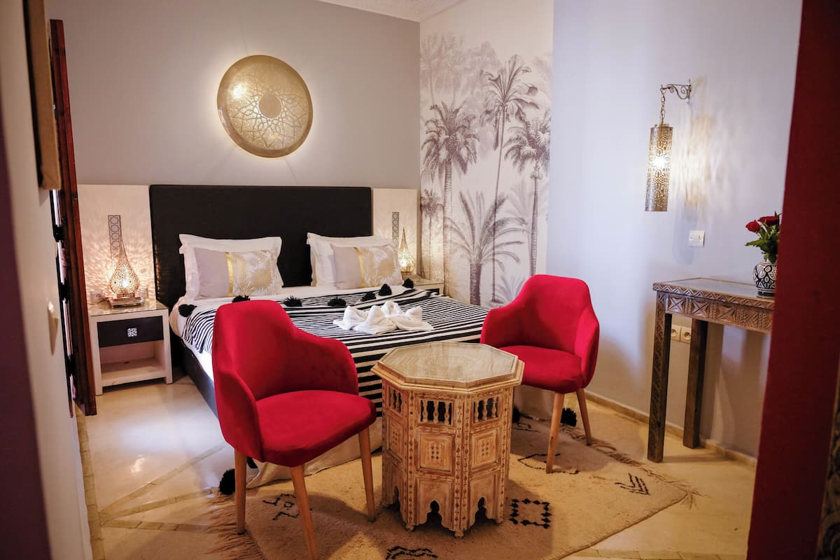 A bedroom with a bed, two red chairs, a small round table, a nightstand, a wall-mounted lamp, and a decorative wall piece.