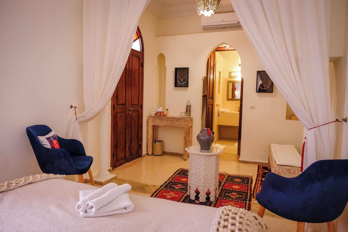 A hotel room with a bed, two blue chairs, a small table with a vase, a mirror, and a doorway leading to another room.