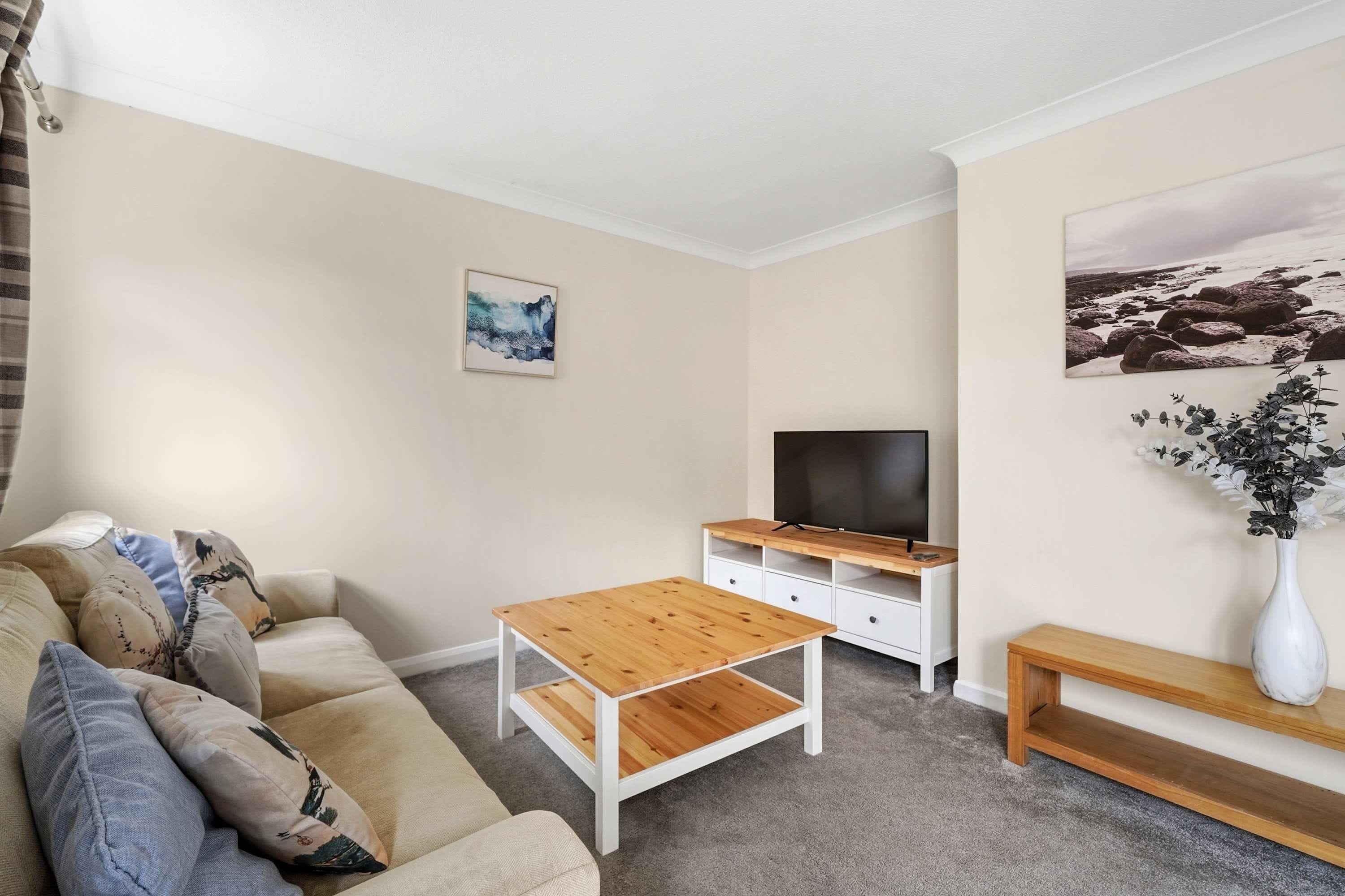 Una sala de estar con un sofá beige, una mesa de centro de madera, un mueble para la televisión y un cuadro en la pared.