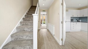 Un escalier menant à une porte, une cuisine avec des meubles blancs et un plan de travail bleu, et un couloir avec un revêtement de sol de couleur claire.