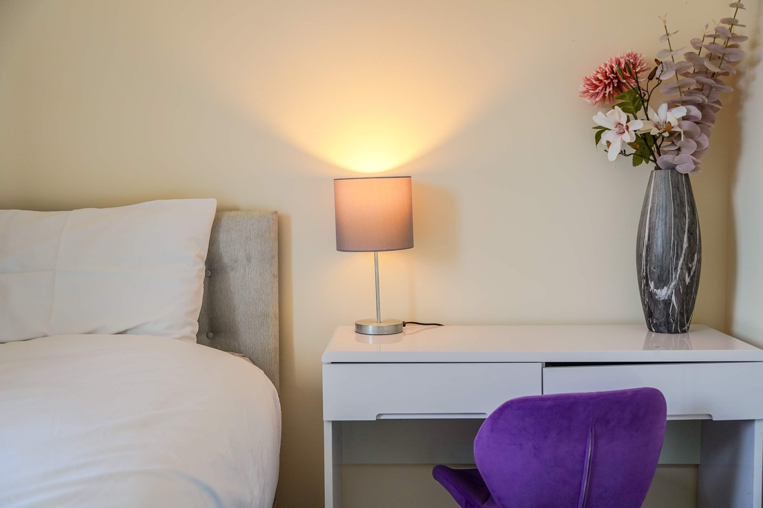 A hotel room with a bed, a desk with a lamp, a purple chair, and a vase with flowers.