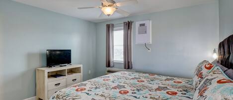 A bedroom with a bed, a ceiling fan, a TV on a stand, a window with curtains, and an air conditioning unit.