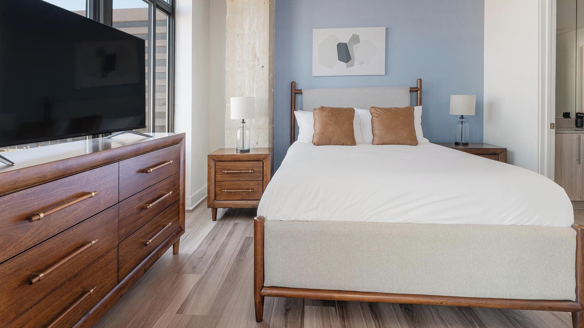 A bedroom with a bed, wooden dresser, nightstands, a television, and a wall-mounted artwork.