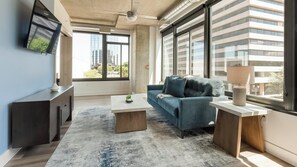 A modern living room with a sofa, coffee table, and TV, large windows with city views, and a ceiling fan. - Landing | Stunning 2BD, Gym, Game Room (Arlington)
