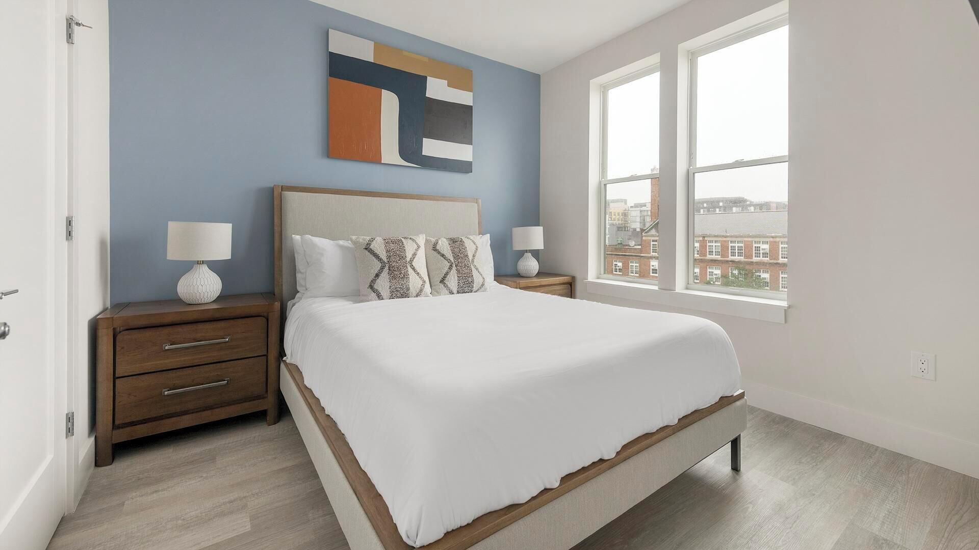 A bedroom with a bed, nightstands, a wall art piece, and a window with a city view.