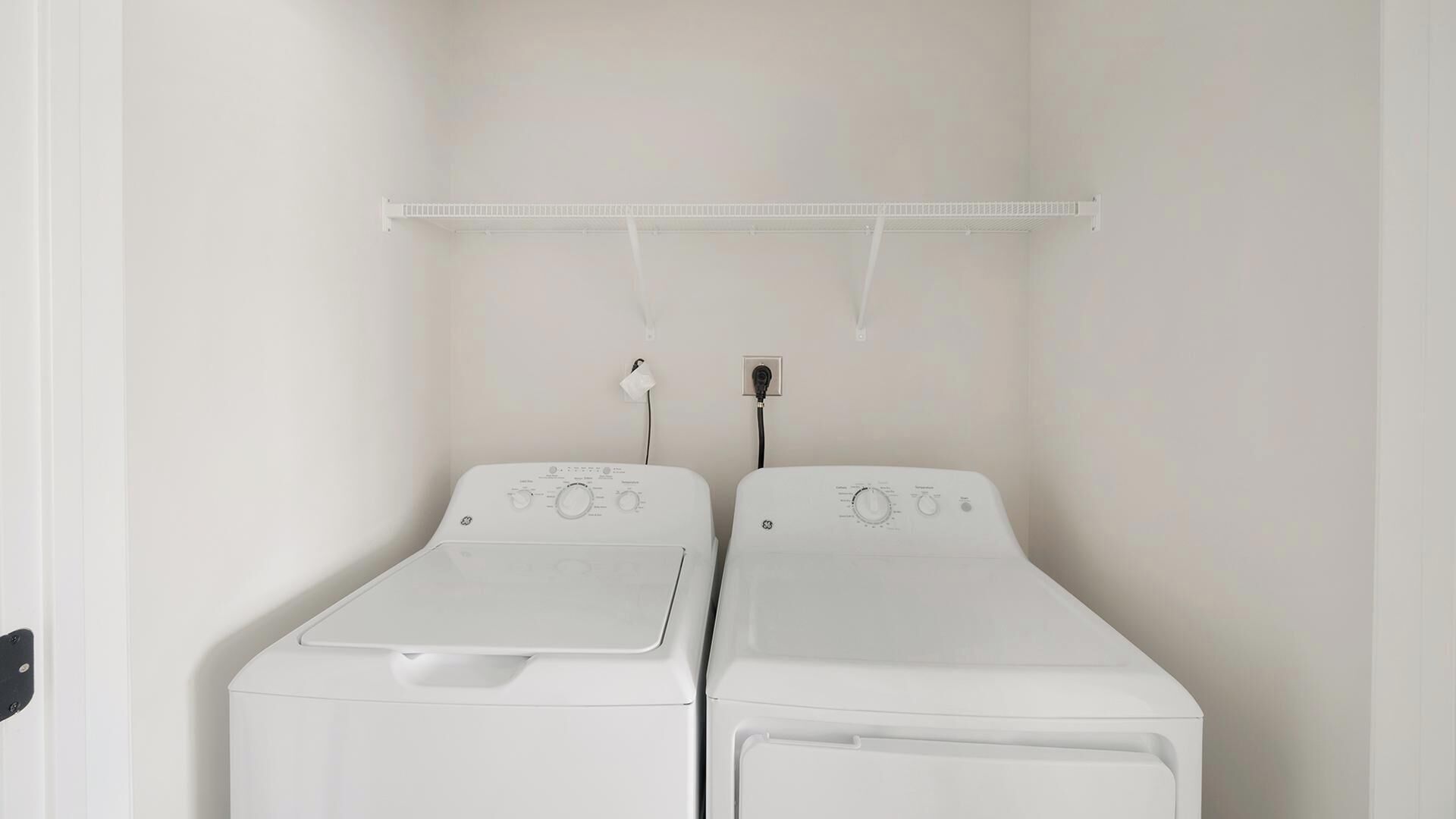 Une buanderie équipée de deux machines à laver à chargement frontal blanches, d’une étagère blanche au-dessus et d’une prise de courant sur le mur.