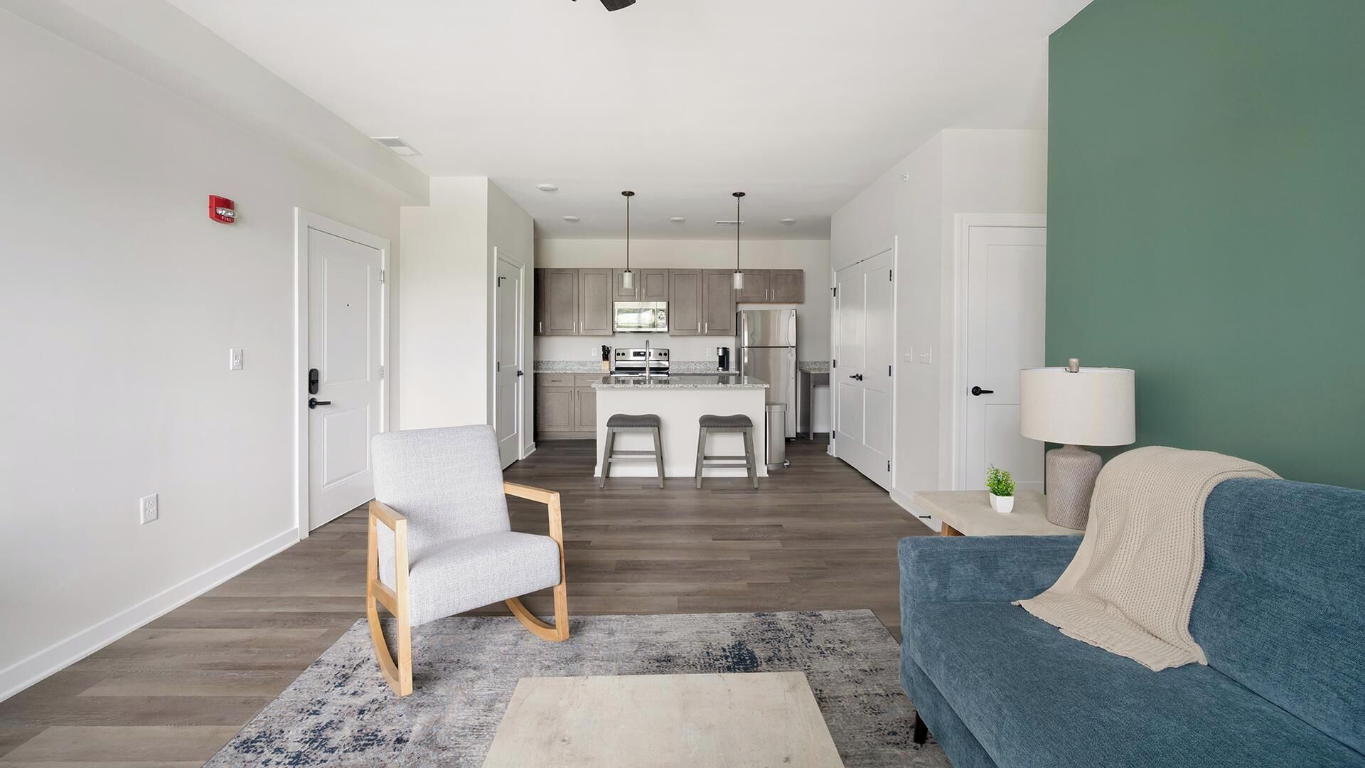 Un apartamento moderno con cocina, sala de estar con sofá azul y sillón gris, y una pequeña mesita de noche con lámpara.
