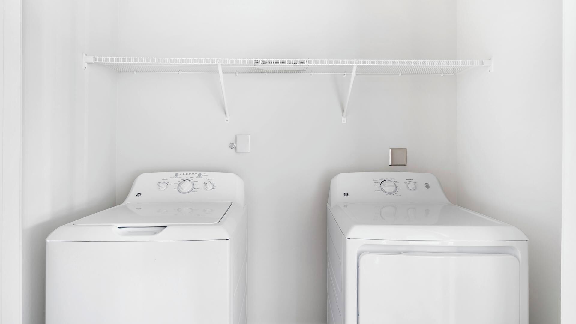 Deux machines à laver à chargement frontal, de couleur blanche, se trouvent dans une buanderie, avec une étagère en métal au-dessus.