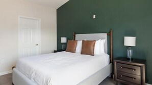 Ein ordentlich bezogenes Bett mit weißer Bettwäsche und zwei braunen Kissen, begleitet von zwei Nachttischen mit Lampen, in einem Zimmer mit grüner Akzentwand und weißer Tür.