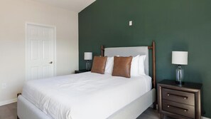 A neatly made bed with white bedding and two brown pillows, flanked by two bedside tables with lamps, in a room with a green accent wall and a white door. - Landing | Stunning 2BD, Gym (Garland)