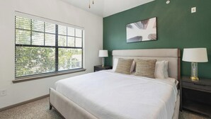 Quarto com uma janela grande, uma cama com roupa de cama branca, duas mesas de cabeceira com abajures, e uma parede verde decorativa.