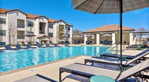 A swimming pool with lounge chairs and umbrellas, surrounded by buildings with a tiled roof. - Landing | Spectacular 2BD, Gym (Garland)