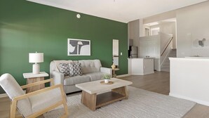 Ein Wohnzimmer mit grüner Wand, einer Couch, einem Couchtisch und einem Sessel. Im Hintergrund ist eine Küchenzeile mit einem Kühlschrank und einer Arbeitsplatte zu sehen.