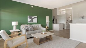A living room with a green wall, a sofa, a coffee table, and a chair. There is a kitchen area with a fridge and a counter visible in the background. - Landing | Amazing 2BD, Gym (Garland)