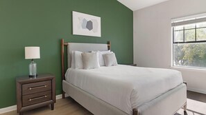 A bedroom with a bed, a nightstand, a lamp, and a window with a view of trees. - Landing | Modern 2BD, Gym (Garland)
