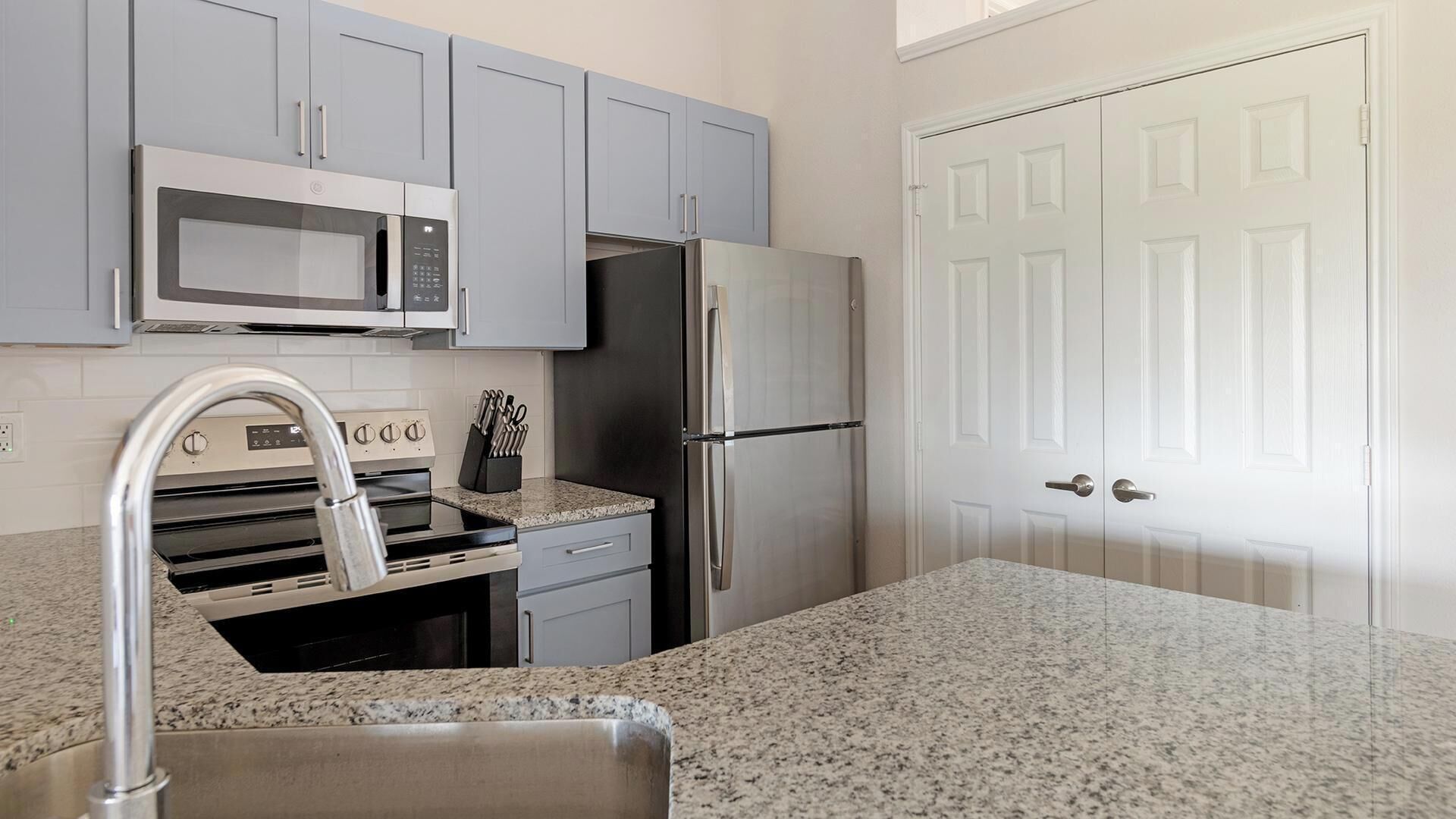 A kitchen with grey cabinets, a stainless steel refrigerator, a microwave, and a granite countertop.