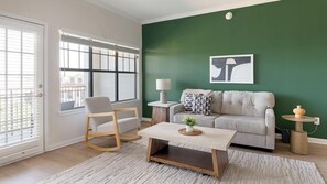 A living room with a green wall, a sofa, a rocking chair, a coffee table, and a window with blinds. - Landing | Modern 2BD, Gym (Garland)