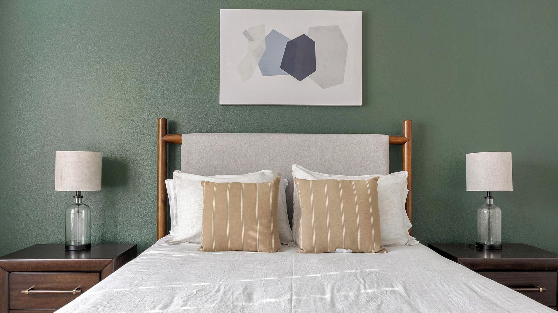 Een net opgemaakt bed met een witte dekbedovertrek, twee nachtkastjes met lampen, en een ingelijst abstract kunstwerk aan de muur.