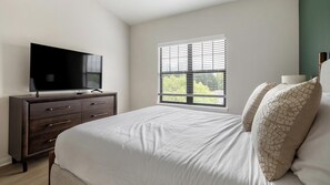 A bedroom with a bed, a dresser, a television, and a window with blinds. - Landing | Chic 2BD, Gym (Garland)