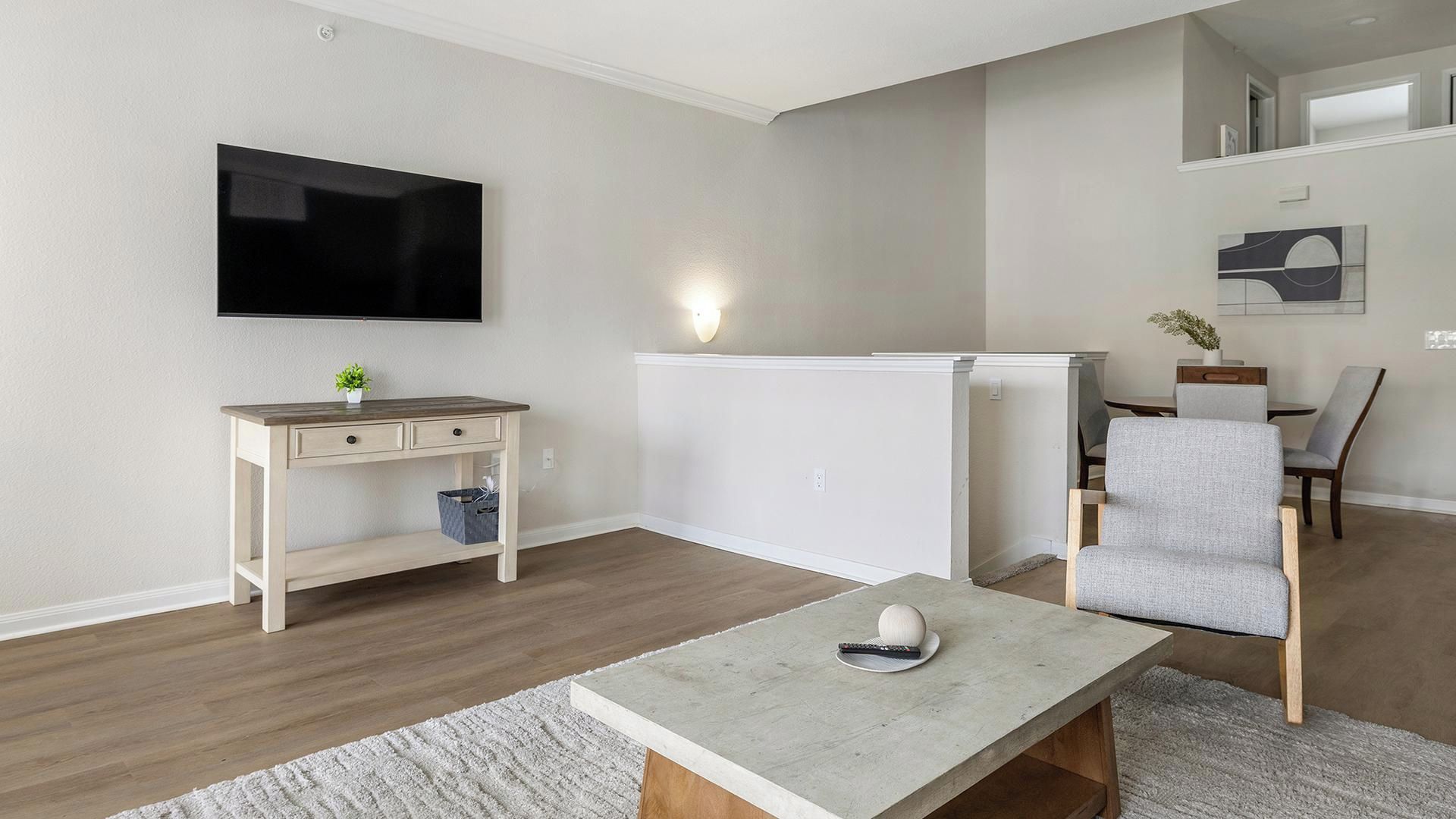 A modern living room with a flat-screen TV mounted on the wall, a wooden desk with drawers, a grey armchair, and a concrete coffee table.
