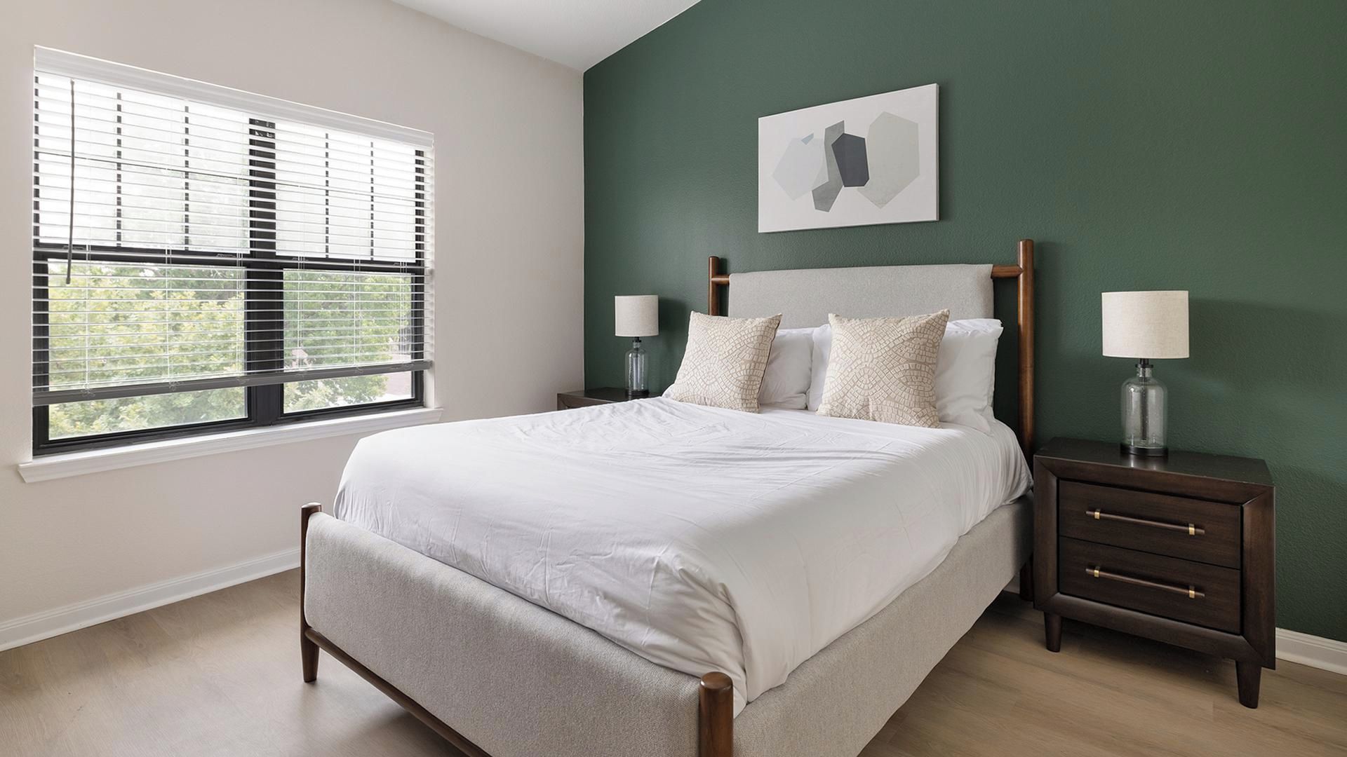 Een slaapkamer met een bed, twee nachtkastjes, een raam met jaloezieën en een wanddecoratie.