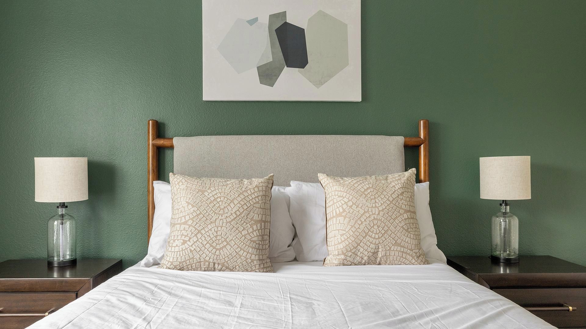 A neatly made bed with a wooden headboard, two bedside lamps, and a wall-mounted abstract art piece.