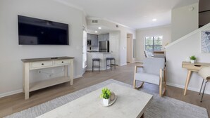 En moderne stue med et fladskærms-TV, et spiseområde med bord og stole, et køkken med bar samt en trappe til en etage over.