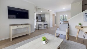A modern living room with a flat-screen TV, a dining area with a table and chairs, a kitchen with a bar, and a staircase leading to an upper level. - Landing | Stylish 2BD, Gym (Garland)