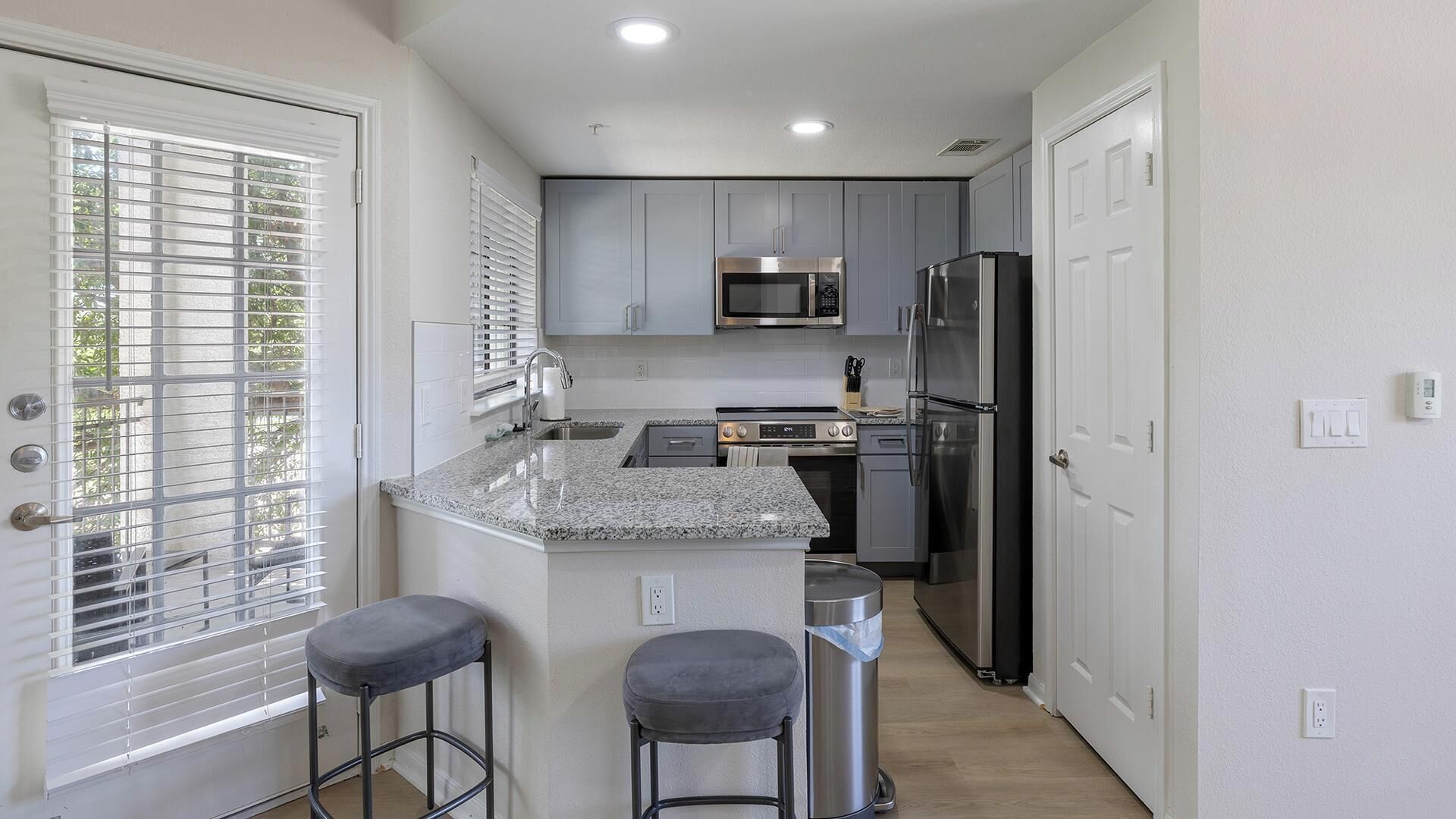 A modern kitchen with grey cabinets, stainless steel appliances, a granite countertop, and two bar stools.
