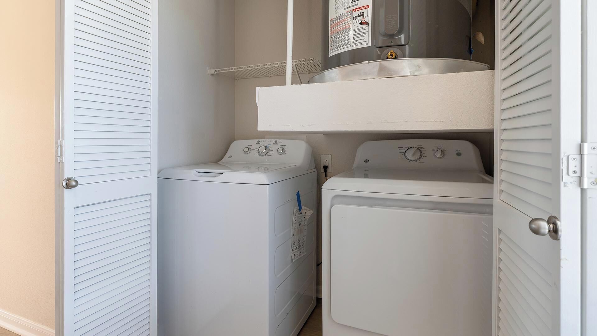 A laundry room with two white dryers, a shelf above, and a door on the left.