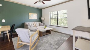 A living room with a sofa, coffee table, and a rocking chair. There's a window with blinds, a ceiling fan, and a painting on the wall.