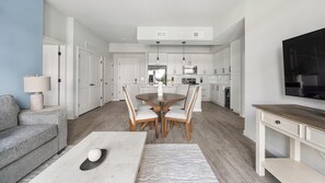 Eine moderne Küche mit weißen Schränken, ein Essbereich mit rundem Tisch und Stühlen, ein graues Sofa und ein flachbildfernseher an der Wand.