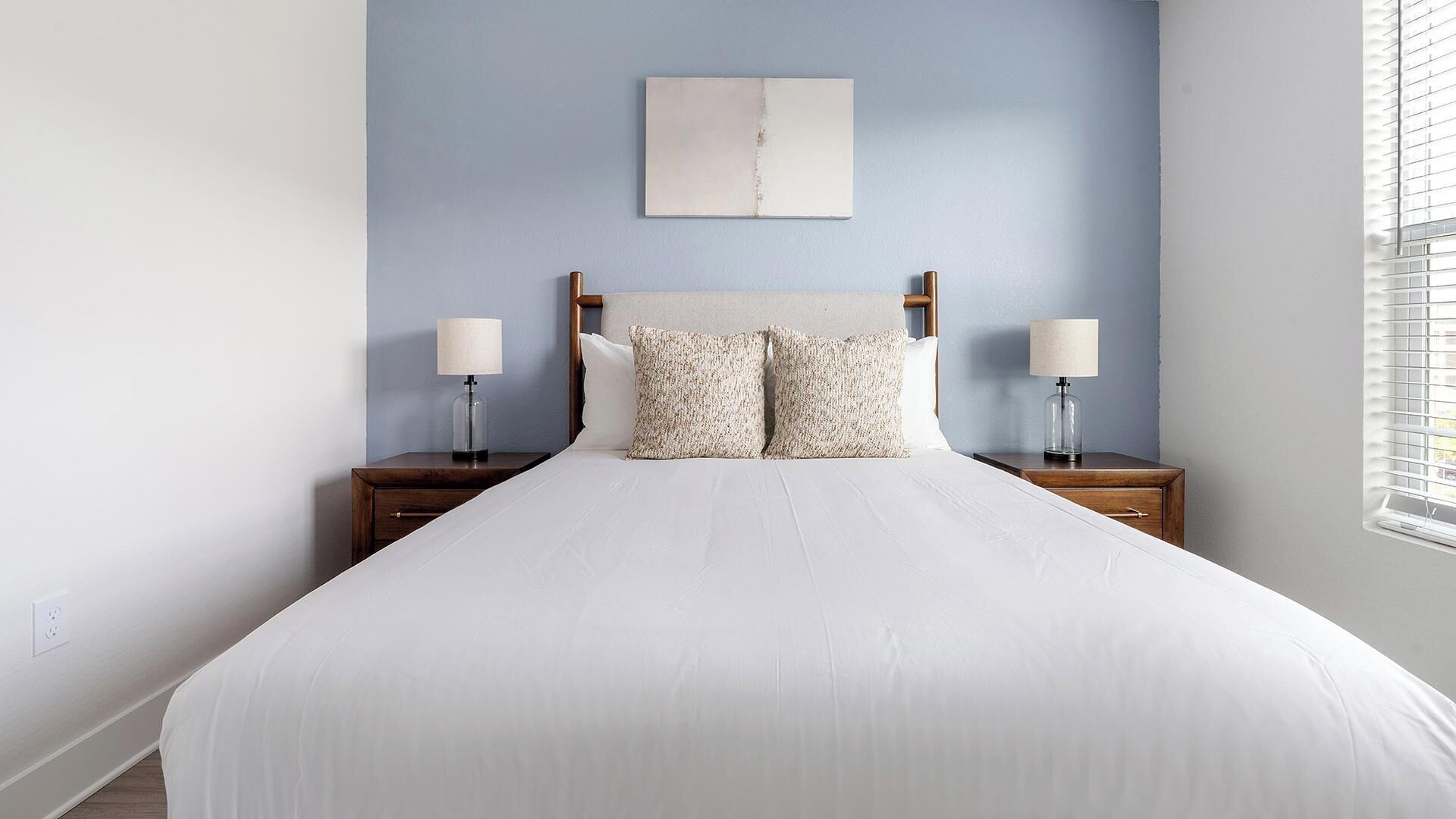 Quarto com cama, duas mesas de cabeceira, luminárias, um criado-mudo, uma obra de arte na parede e uma janela com persianas.