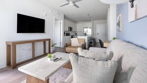 Ein modernes Wohnzimmer mit einem Flachbildfernseher, einem Holztisch, einem beigefarbenen Sofa und einem Essbereich im Hintergrund.