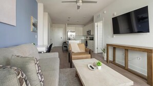 Una sala de estar moderna con un sofá, una mesa baja con una planta, un televisor montado en la pared y una cocina al fondo.